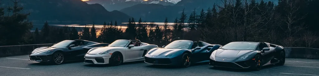 A line of high-performance luxury convertible supercars, including a Chevrolet Corvette, Porsche 718 Spyder, Ferrari 488 Spider, and Lamborghini Huracán, parked at a scenic mountain overlook during sunset; provided by Starr Luxury.