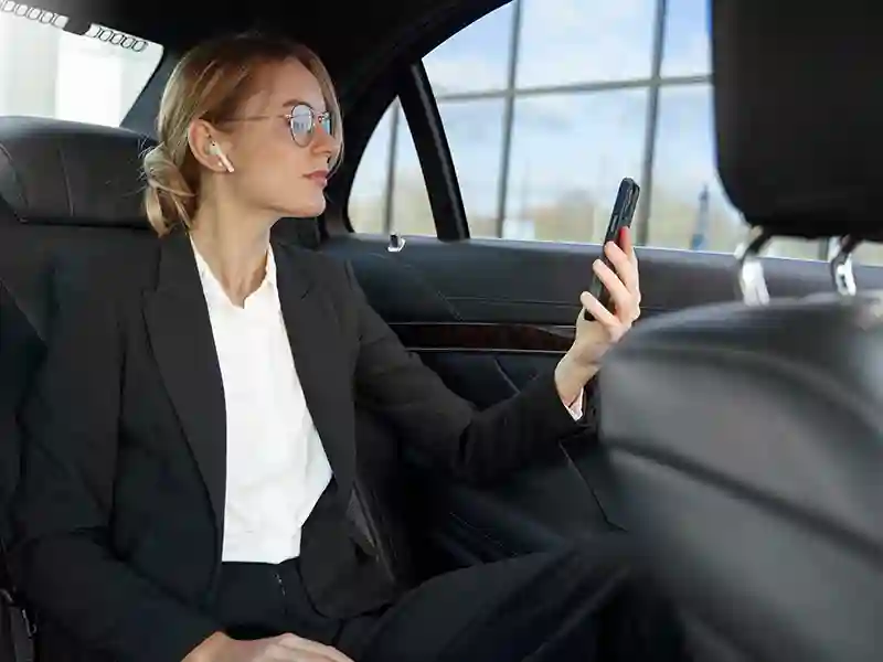 Inside the back seat, a blond woman with glasses in a grey suit is reading a message on her mobile