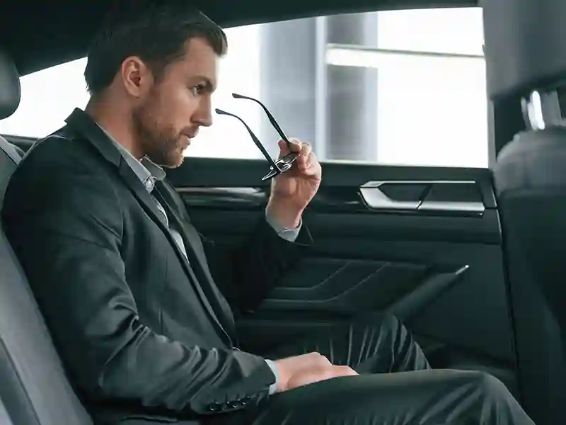 A man inside of a car in the back seat, wearing his sunglasses