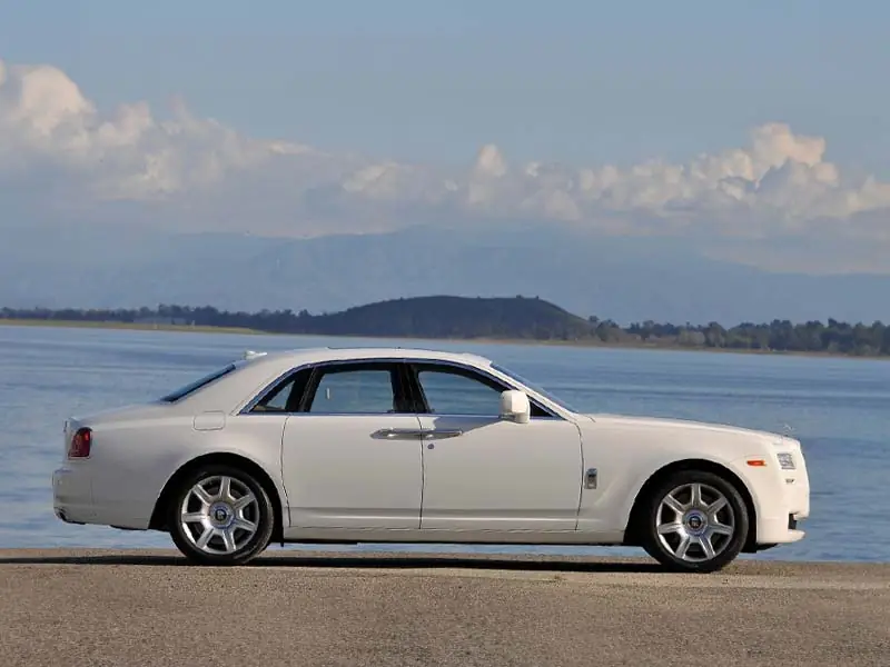 A sleek grey Rolls-Royce Ghost driving through a modern financial district, featuring blacked-out wheels, the signature chrome Pantheon grille, and Starr Luxury branding.