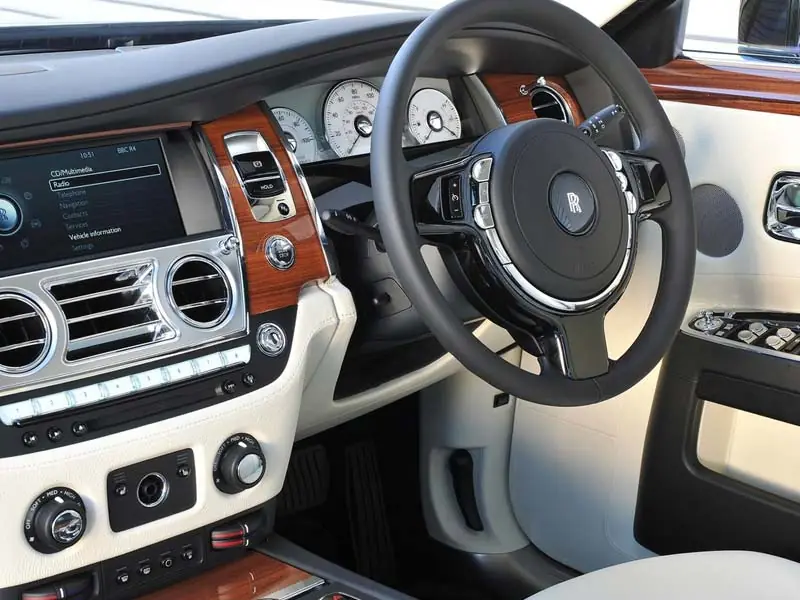 The front cabin of a Rolls-Royce, featuring a white leather dashboard accented by warm teak wood veneer, a digital infotainment screen, chrome air vents, and a multi-function steering wheel with the RR emblem.