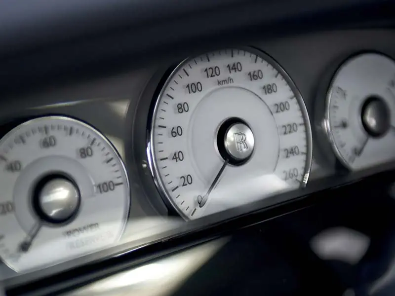 A close-up of the classic Rolls-Royce Ghost instrument dials, featuring a white-faced speedometer and the iconic "Power Reserve" gauge with the RR logo at the center.
