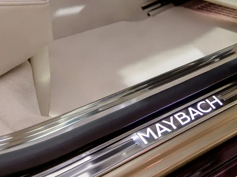 A close-up of an illuminated chrome door sill featuring the "MAYBACH" logo in bright white light, set against premium cream-colored plush carpeting in a luxury sedan.