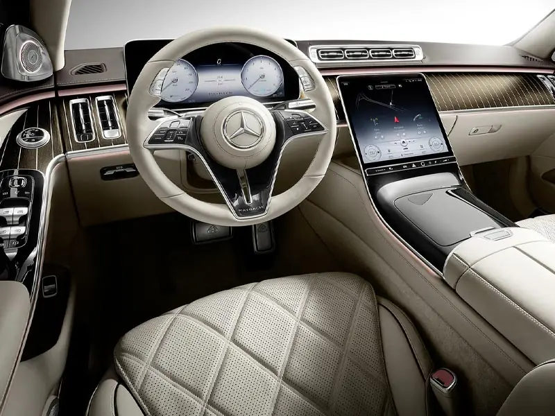 A wide interior view of a Mercedes-Maybach cockpit featuring a cream-colored leather steering wheel, quilted leather seats, pinstriped wood trim, and a high-resolution dual-screen digital dashboard.