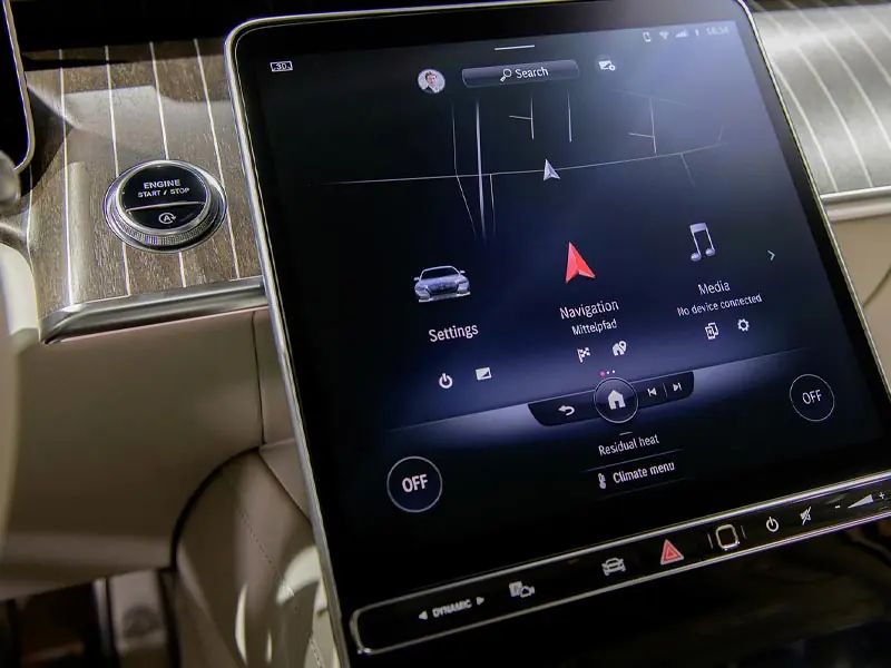 A close-up of a large vertical touchscreen infotainment system in a Mercedes-Maybach, displaying navigation and media settings next to a pinstriped wood dashboard and a silver engine start button.