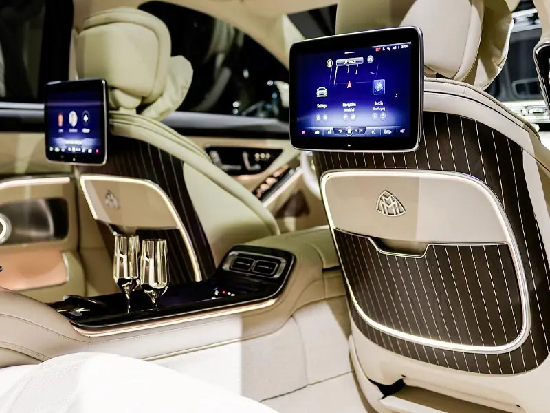 The interior rear cabin of a Mercedes-Maybach featuring cream leather seats, pinstriped wood accents, dual entertainment screens, and two silver champagne flutes set in a center console.