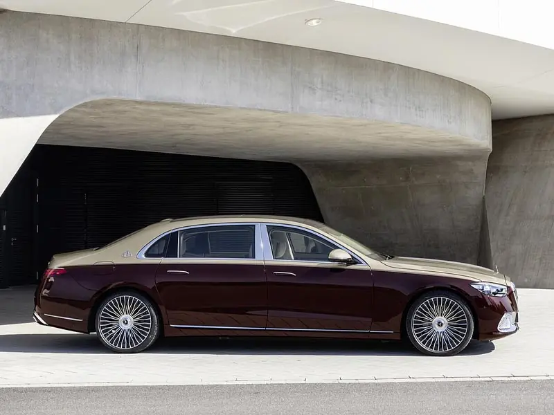 A side-view profile of a two-tone gold and deep burgundy Mercedes-Maybach S-Class parked in front of a modern, minimalist concrete building, showcasing its multi-spoke luxury wheels and extended wheelbase.