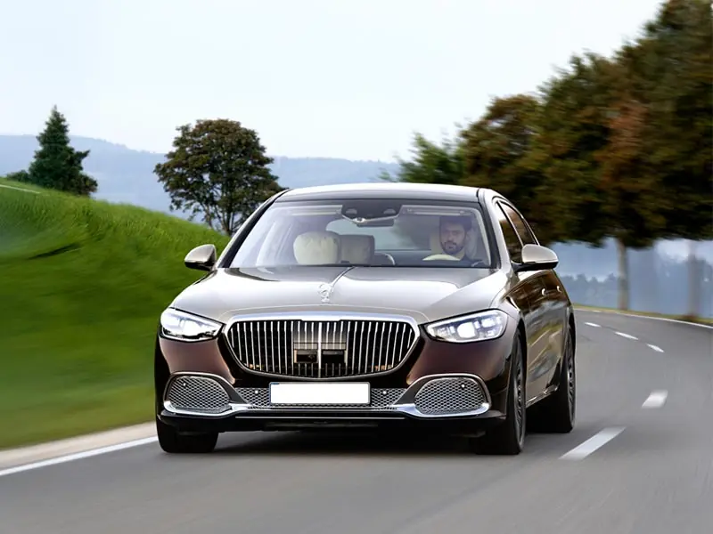 A high-end two-tone Mercedes-Maybach luxury sedan driving along a scenic winding road, featuring a prominent vertical-slat chrome grille and sophisticated LED headlights.