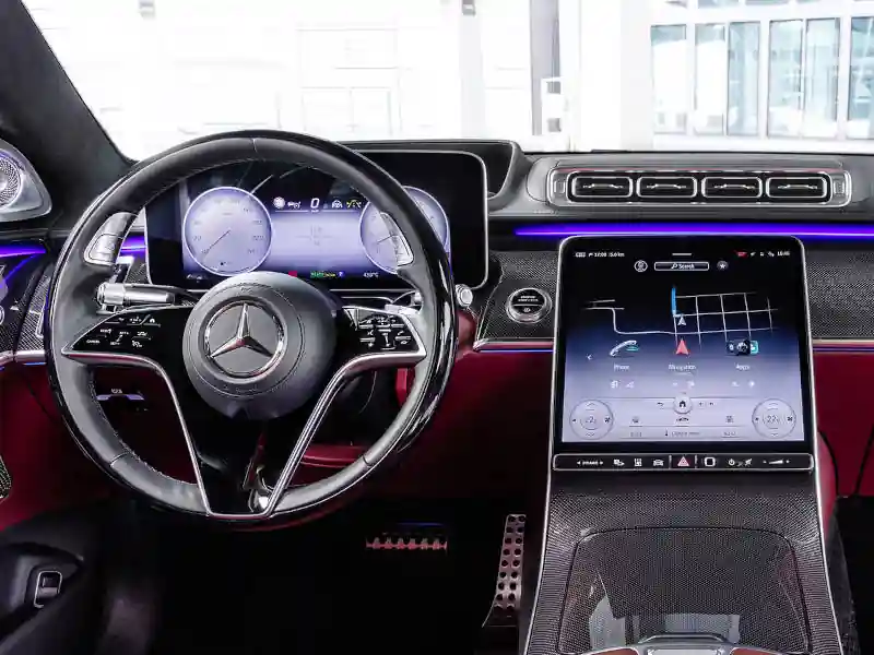 Interior view of a Mercedes-Benz S-Class cockpit featuring deep red leather upholstery, carbon fiber trim, a leather-wrapped steering wheel, and a large vertical touchscreen navigation system with vibrant purple ambient lighting.
