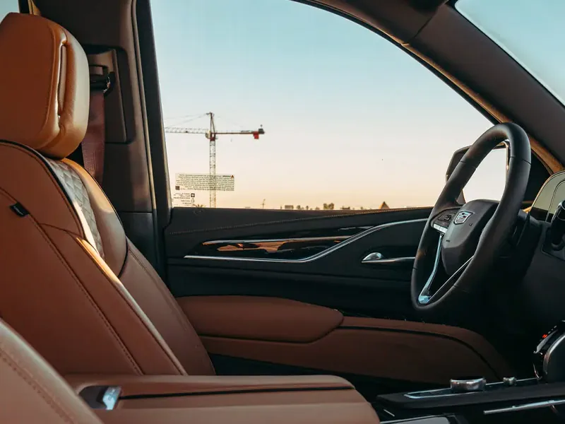 Interior view from the passenger side of a luxury Cadillac, showing tan leather quilted seats and a sleek dashboard, with a city skyline and a construction crane visible through the window during golden hour.