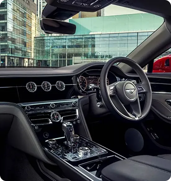 A view from the passenger side of a Bentley Continental GT interior, showcasing the handcrafted leather dashboard, iconic rotating display with three analog dials, and a sleek center console, set against a backdrop of modern glass city buildings.