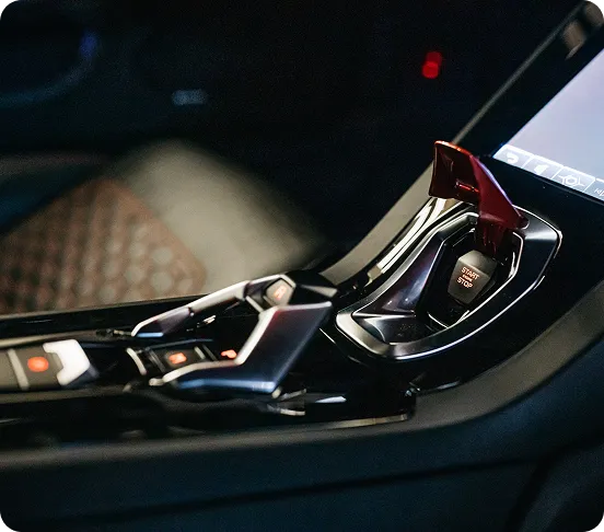 A close-up shot of a Lamborghini Aventador's center console, highlighting the iconic fighter-jet style red flip cover over the engine start-stop button, alongside the metallic gear selector switches.